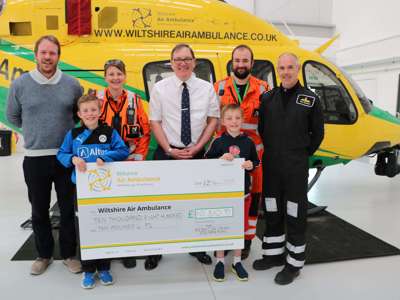 A group of visitors in front of the helicopter holding a large cheque inside the hanger A group of visitors in front of the helicopter holding a large cheque inside the hanger