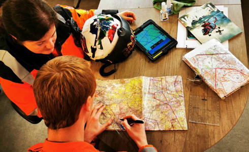 Two paramedics who are both wearing orange flight suits. They are both sat at a wooden desk with an iPad and paper maps and are plotting a route. Two paramedics who are both wearing orange flight suits. They are both sat at a wooden desk with an iPad and paper maps and are plotting a route.