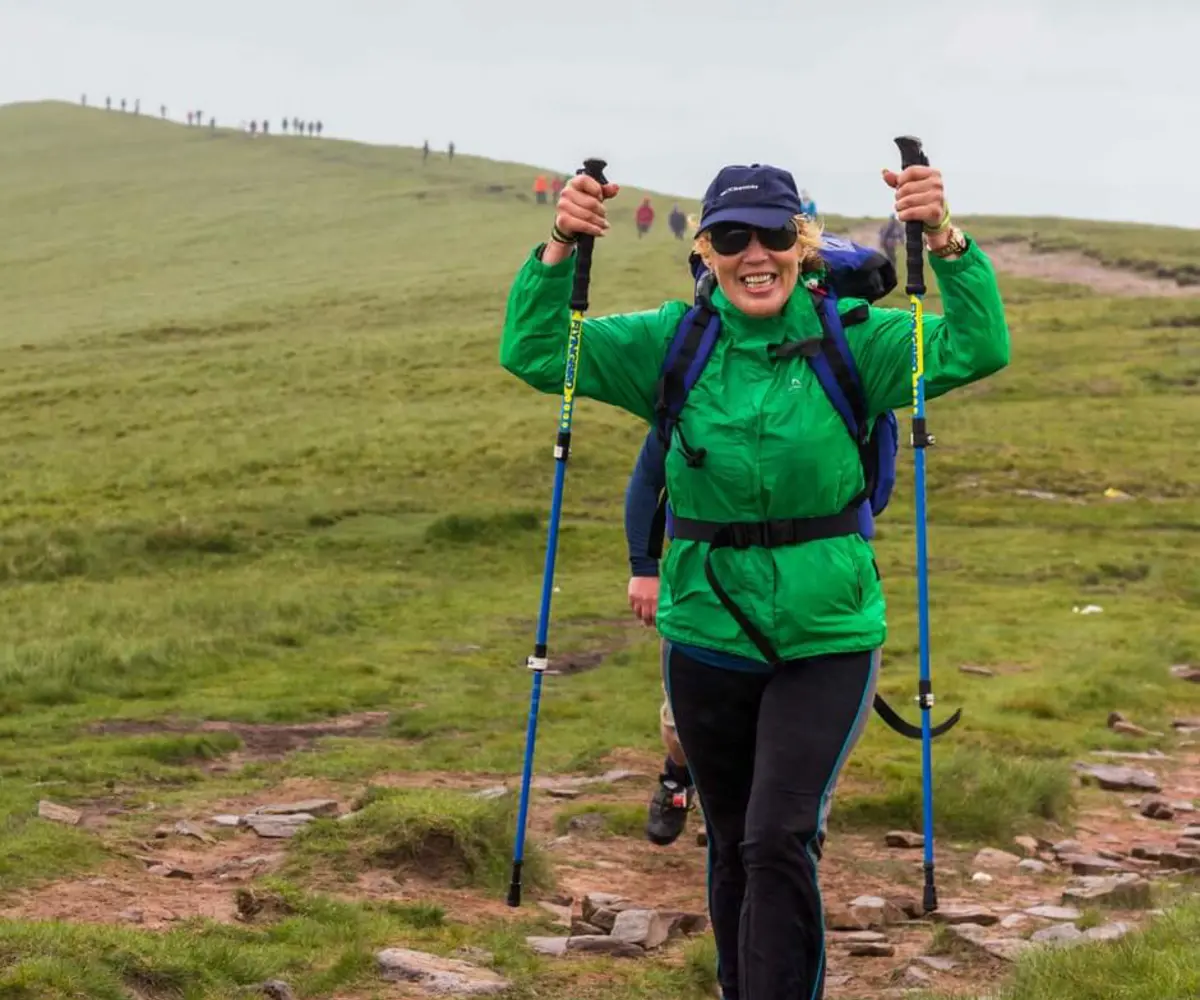 A person wearing a green raincoat and blue backpack, holding walking poles in the air