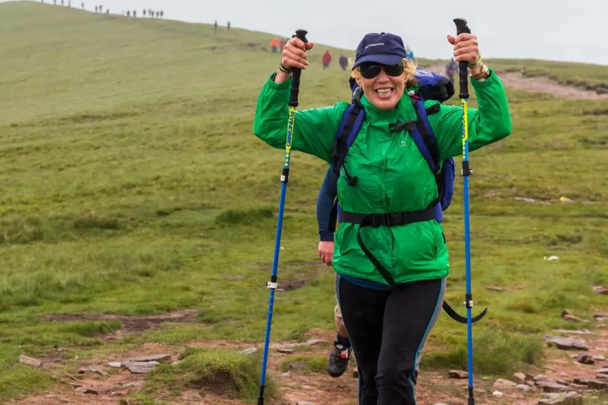 A person wearing a green raincoat and blue backpack, holding walking poles in the air