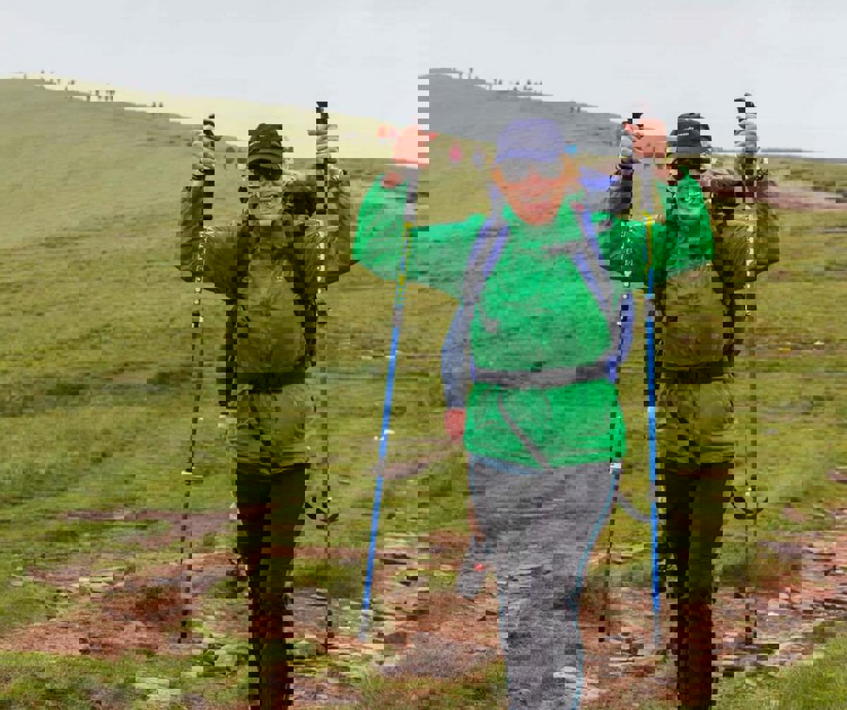 A person wearing a green raincoat and blue backpack, holding walking poles in the air