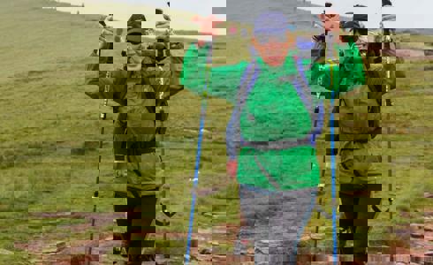 A person wearing a green raincoat and blue backpack, holding walking poles in the air A person wearing a green raincoat and blue backpack, holding walking poles in the air