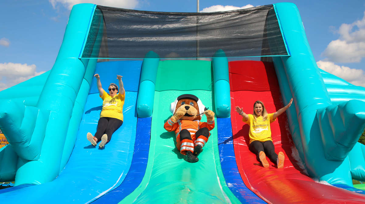 Wilber, bear mascot, sliding down an inflatable slide with two women wearing yellow Wiltshire Air Ambulance t-shirts Wilber, bear mascot, sliding down an inflatable slide with two women wearing yellow Wiltshire Air Ambulance t-shirts