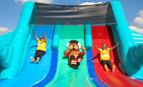 Wilber, bear mascot, sliding down an inflatable slide with two women wearing yellow Wiltshire Air Ambulance t-shirts Wilber, bear mascot, sliding down an inflatable slide with two women wearing yellow Wiltshire Air Ambulance t-shirts