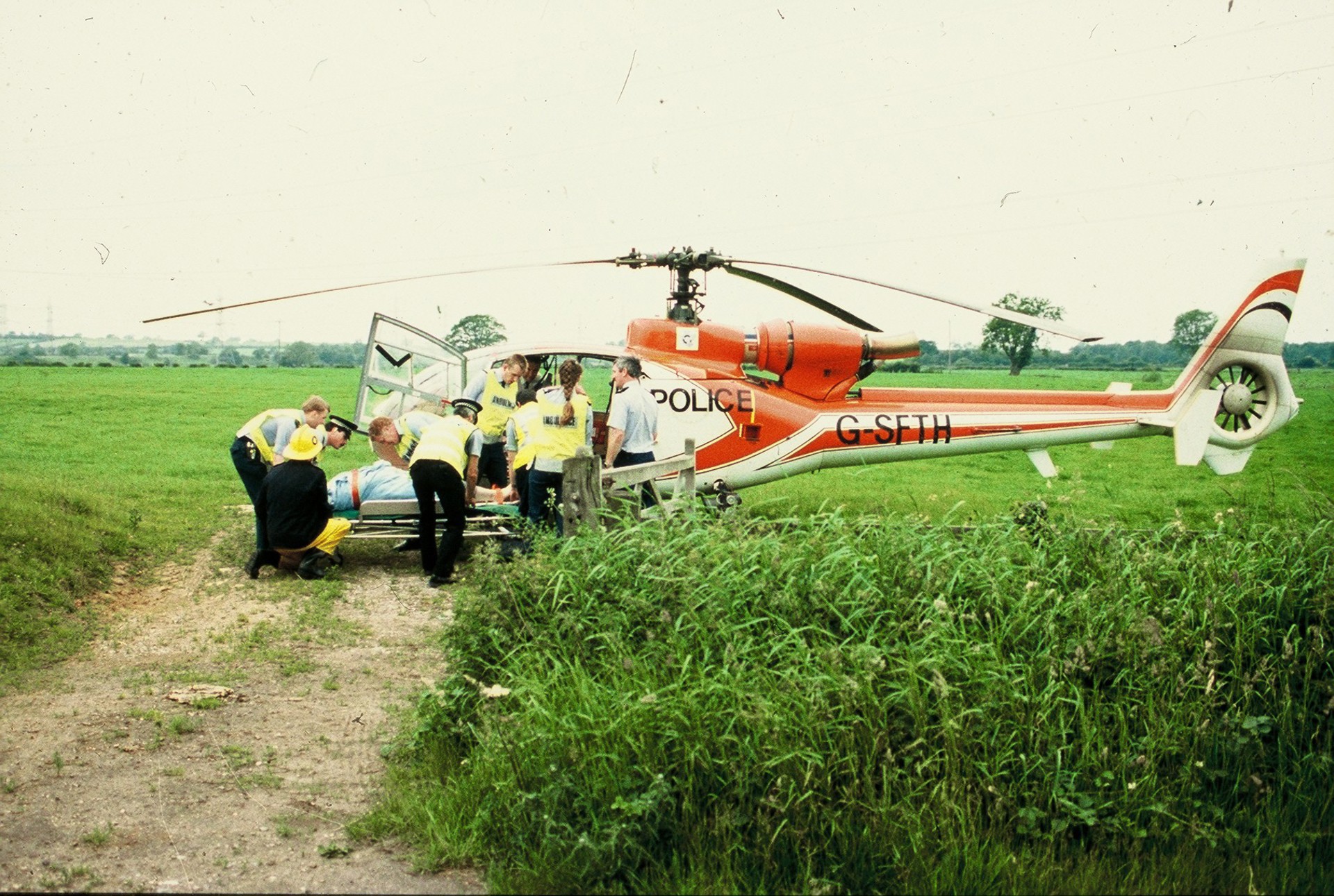 A scanned photo of a red helicopter landed in a field with police lifting a patient on a stretcher.