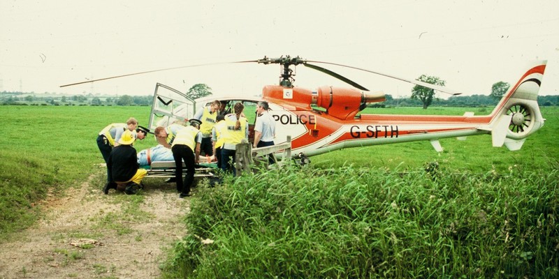 A scanned photo of a red helicopter landed in a field with police lifting a patient on a stretcher. A scanned photo of a red helicopter landed in a field with police lifting a patient on a stretcher.