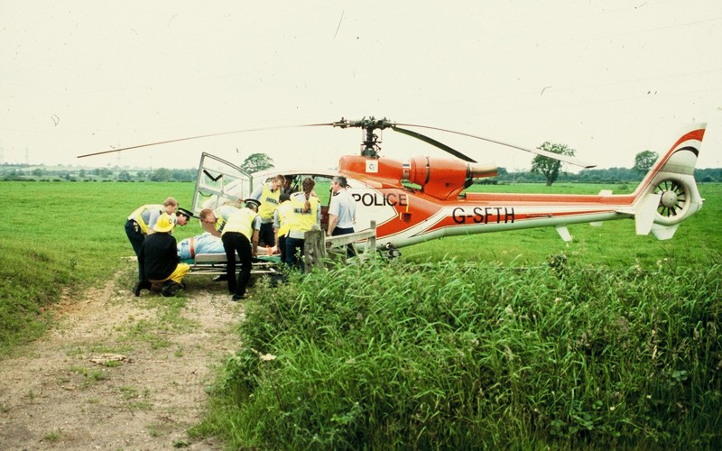 A scanned photo of a red helicopter landed in a field with police lifting a patient on a stretcher. A scanned photo of a red helicopter landed in a field with police lifting a patient on a stretcher.