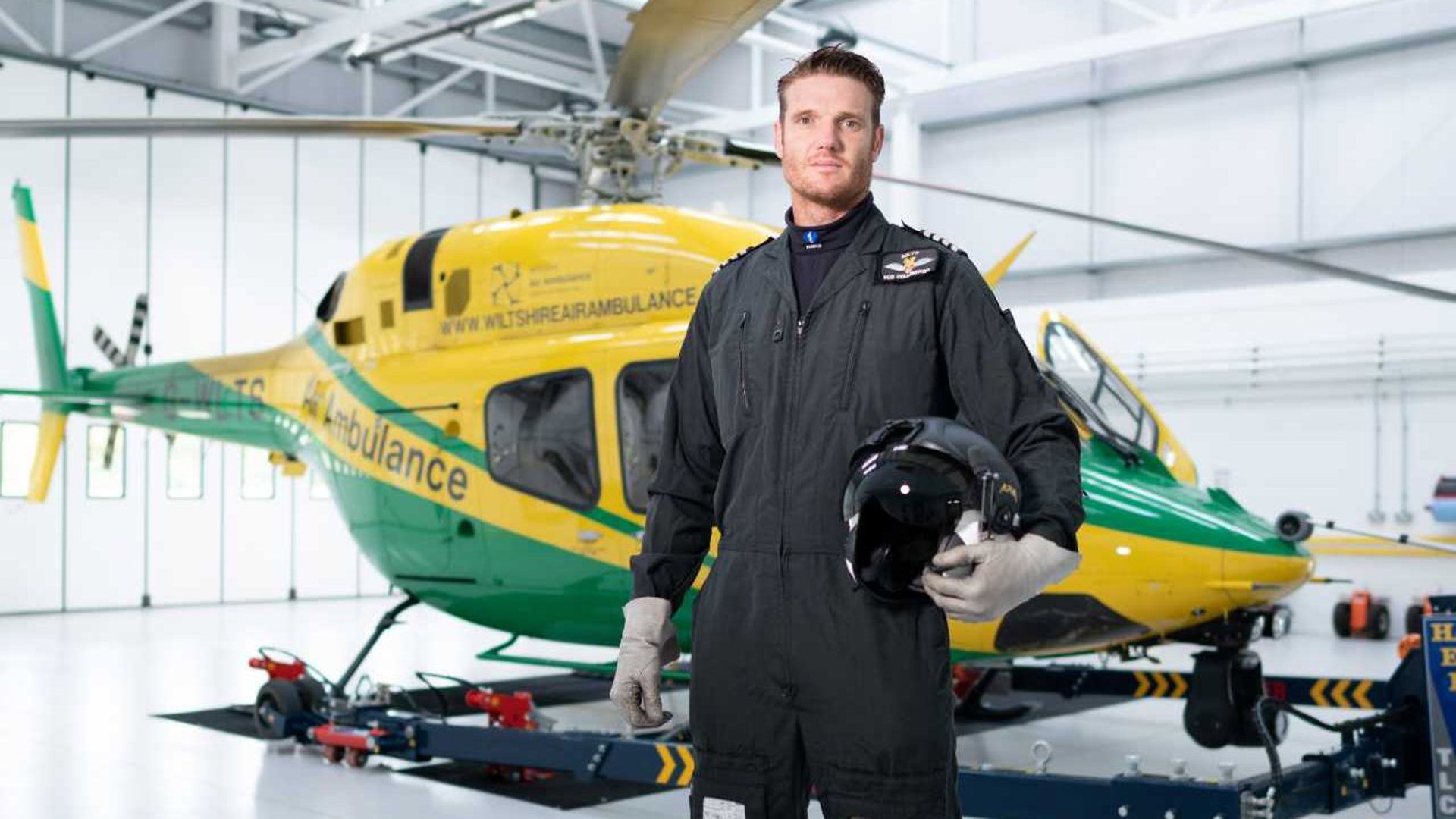 Pilot Rob Collingwood stood wearing a flight suit and holding a flight helmet in front of the helicopter in the hangar. Pilot Rob Collingwood stood wearing a flight suit and holding a flight helmet in front of the helicopter in the hangar.