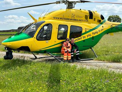 A photo of a paramedic and pilot smiling at the camera whilst perching on the step of the Wiltshire Air Ambulance's yellow and green helicopter A photo of a paramedic and pilot smiling at the camera whilst perching on the step of the Wiltshire Air Ambulance's yellow and green helicopter