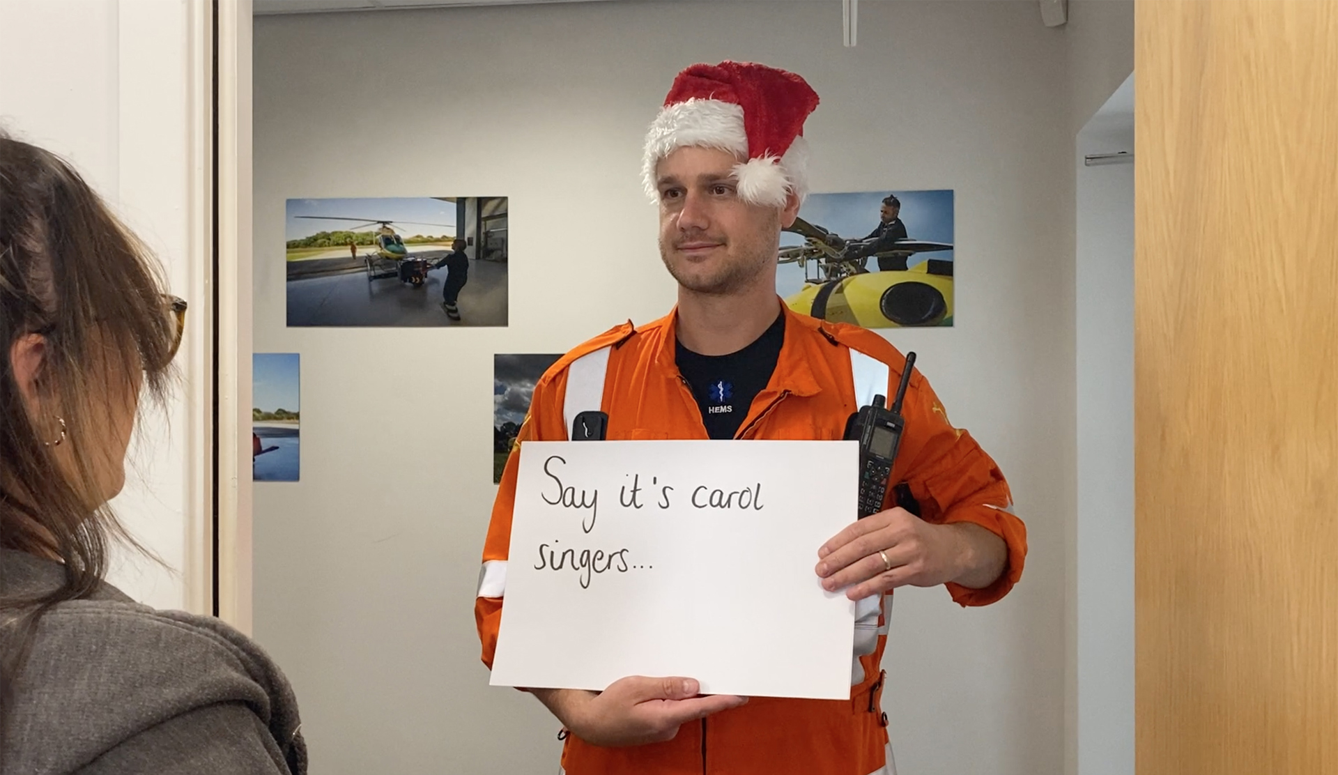 A paramedic wearing a santa hat, holding a "say it's carol singers" sign A paramedic wearing a santa hat, holding a "say it's carol singers" sign